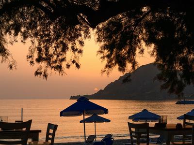 Plaża z stołami, krzesłami i niebieskimi parasolami o zachodzie słońca z wzgórzem w tle.