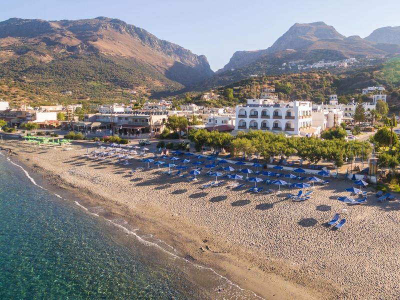 Plage de sable avec parasols et bâtiments devant un paysage montagneux par temps ensoleillé.