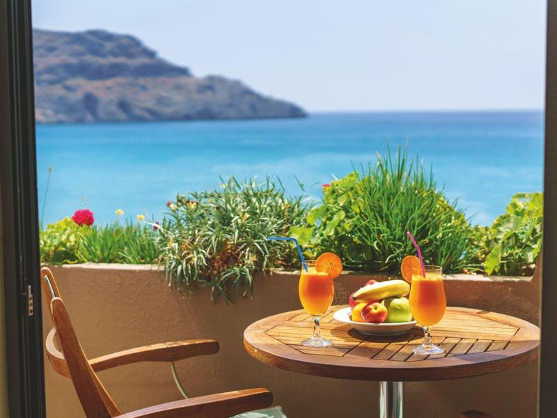 Table de balcon avec boissons et fruits face à la mer et aux montagnes sous un ciel clair.