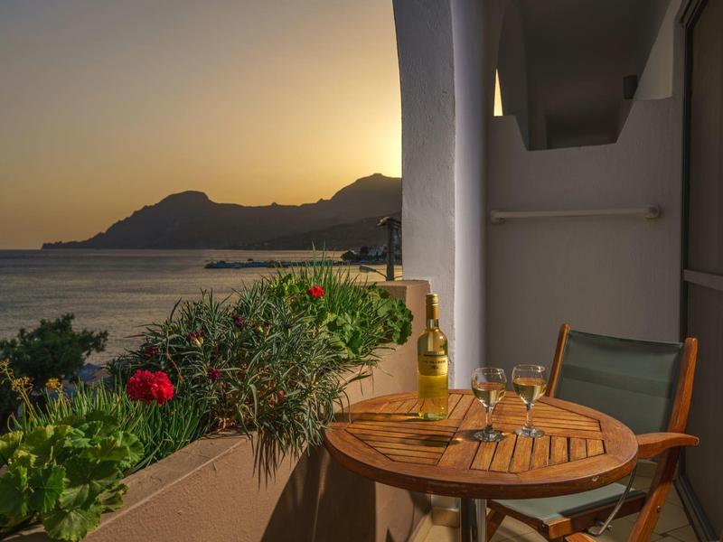 Balcon avec table en bois, chaises et vin au coucher du soleil au bord de la mer avec montagnes en arrière-plan.
