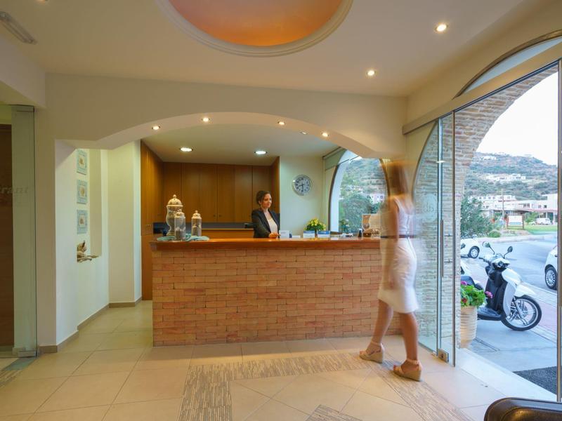 Réception d'hôtel avec réceptionniste derrière un comptoir en briques et une femme en robe blanche marchant à côté.