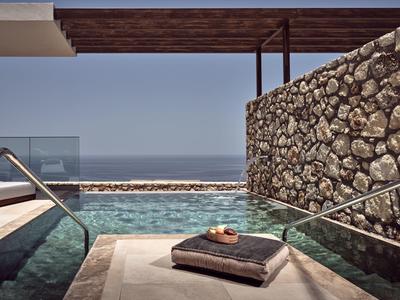 Moderner Poolbereich mit Steinmauer, Glasgeländer, Liegekissen und Blick auf das Meer bei klarem Himmel.