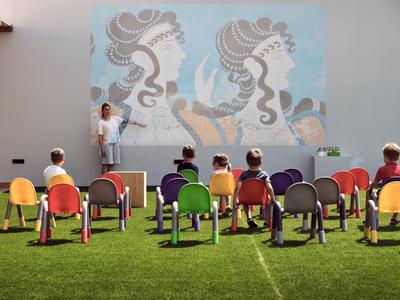 Kinder sitzen auf bunten Stühlen im Freien vor einer Wand mit großem, buntem Wandbild.