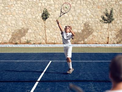 Ein Junge in weißer Kleidung spielt Tennis auf einem blauen Platz mit Steinwand im Hintergrund.