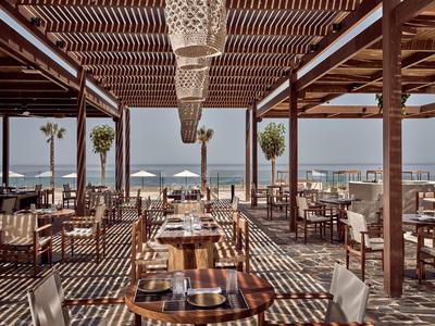 Offenes Restaurant mit Holzdach, Tischen und Stühlen, Blick auf Meer und Sonnenschirme.