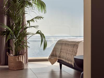 Modernes Schlafzimmer mit Meeresblick, großem Fenster, Palmenpflanze in Korb und leichtem Tageslicht.