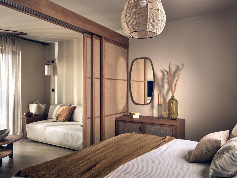Cozy bedroom with wooden accents, beige walls, and a living area visible through sliding doors.