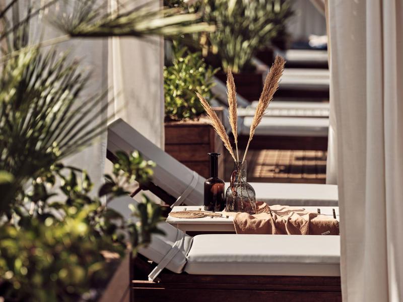 Outdoor lounge area with white cushioned seats, curtains, and wooden decking surrounded by plants.