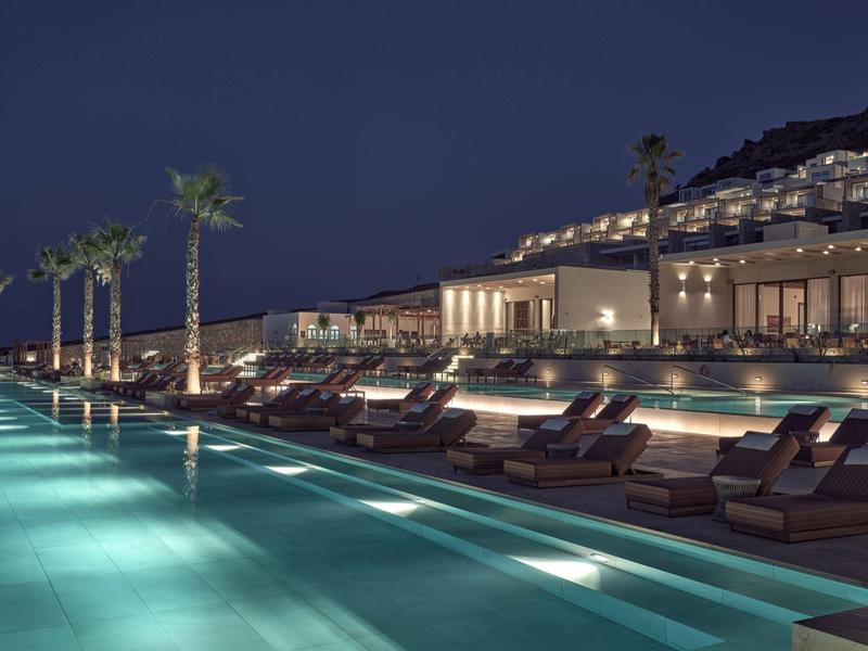 Night view of a lit outdoor pool with sunbeds and palm trees at a hillside hotel.