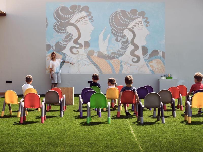 Children seated on colorful chairs watching a puppet show with painted backdrop outdoors.