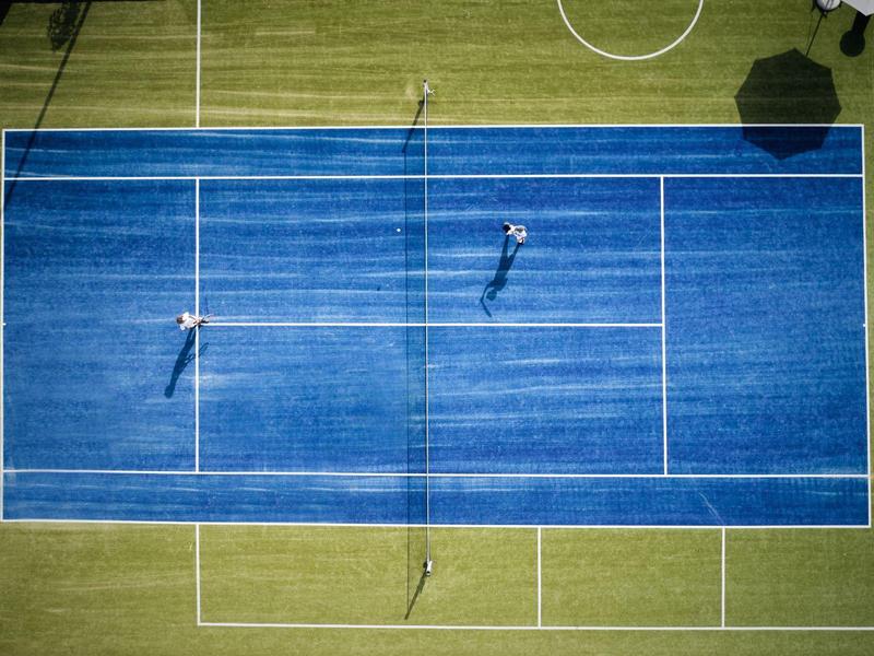 Aerial view of two players on a blue tennis court with green surroundings.