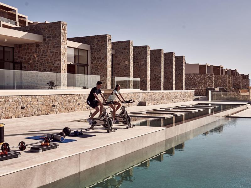 Couple cycling on a pathway beside a pool with stone buildings in the background.
