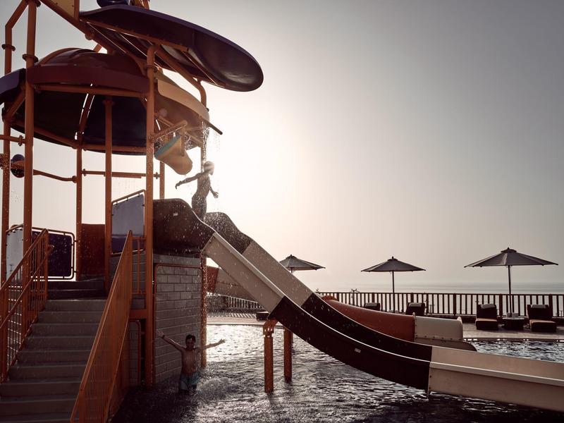 Water slide with sunlit lounge chairs and umbrellas at a beachfront hotel pool.