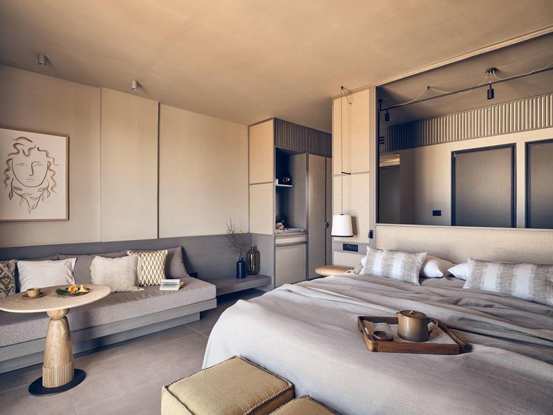 Modern hotel room with double bed, beige decor, and a cozy seating area with cushions.