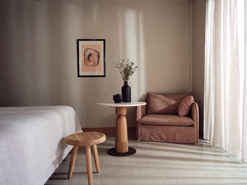 Cozy hotel room with a bed, wooden stool, round table, leather chair, and soft natural light.