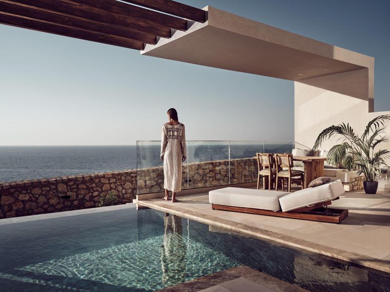 Woman standing by an infinity pool with ocean view under a shaded patio with lounge furniture.