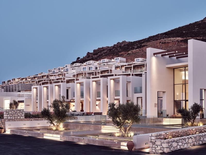Modern white hotel suites with terraces and outdoor pool against hillside at dusk.