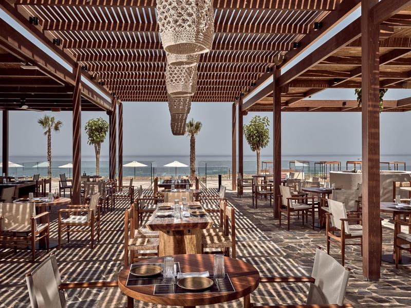Beachside restaurant with wooden tables and chairs under a shaded pergola overlooking the ocean.