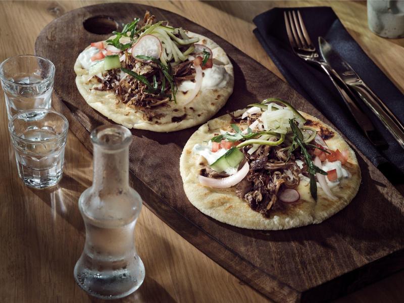 Two flatbreads with meat, olives, and sliced onions on a wooden board, with glasses and utensils nearby.