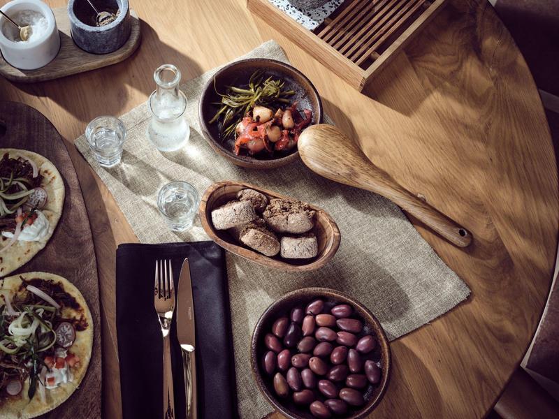 Wooden table with tacos, olives, bread, utensils, and condiments on a neutral cloth.