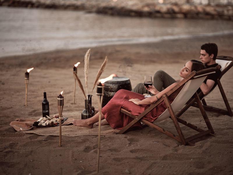 Couple relaxes on beach chairs by torches and wine bottles on sandy shore.