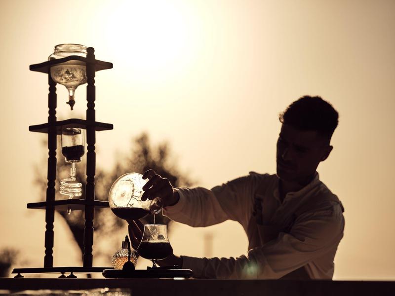 Silhouette of a man pouring coffee from a glass pot outdoors during sunset.