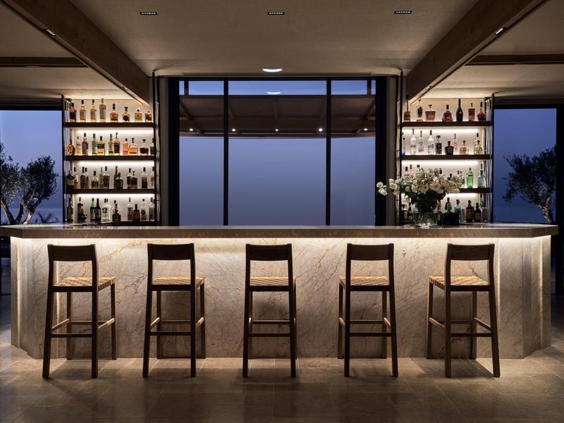 Modern hotel bar at dusk with illuminated shelves and five wooden stools.