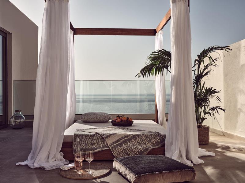 Outdoor canopy bed with white curtains near glass railing and potted plants under clear sky.