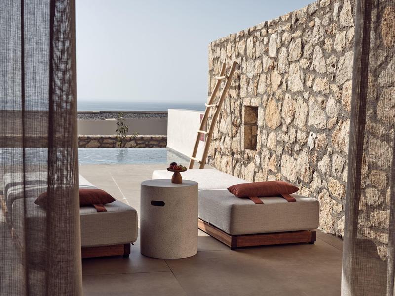 Outdoor lounge area with beige cushions and stone wall near pool and sea view.
