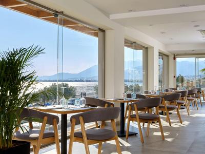 Helles Restaurant mit Holztischen, großen Fenstern und Blick auf Berge und Meer.