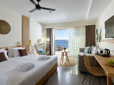 Modernes Hotelzimmer mit Doppelbett, Sitzbereich, Schreibtisch, TV und Meerblick durch Glasfenster.