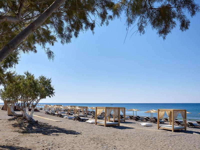 Pláž s lehátky, zastíněnými kabinami a stromy podél pobřeží pod jasnou modrou oblohou.