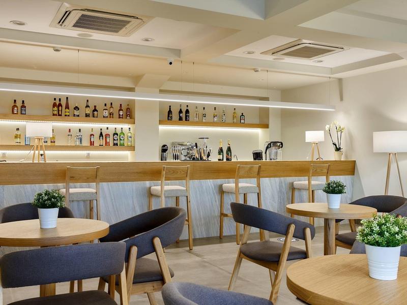 Moderní hotelový salonek s kulatými dřevěnými stoly, šedými židlemi a dobře osvětleným barem.