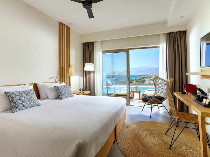 Světý hotelový pokoj s manželskou postelí, balkonem, výhledem na moře, dřevěným stolem a moderním interiérem.