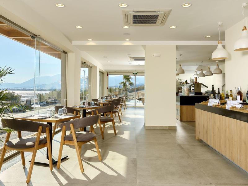 Světelná hotelová restaurace s dřevěnými stolky, židlemi a velkými okny s výhledem na moře.