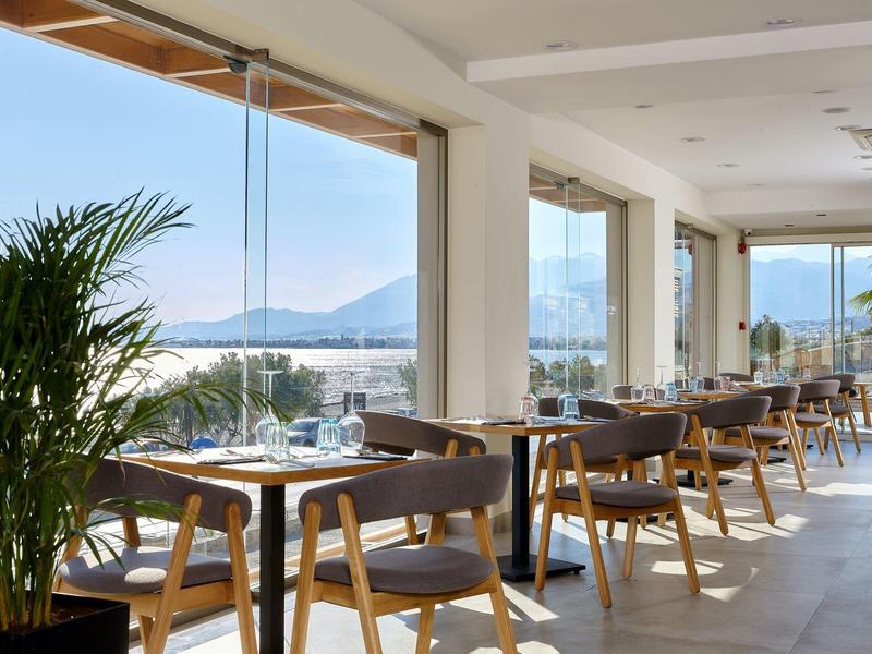 Světá jídelna s dřevěnými stolky a židlemi s výhledem na moře přes velká skleněná okna.