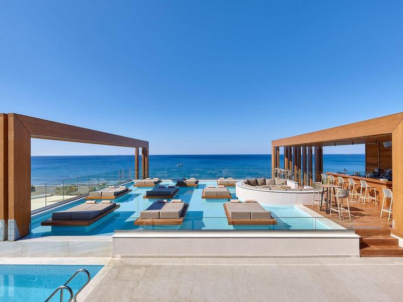 Moderní bazén s dřevěnými pergolami, lehátky a výhledem na oceán pod jasnou modrou oblohou.