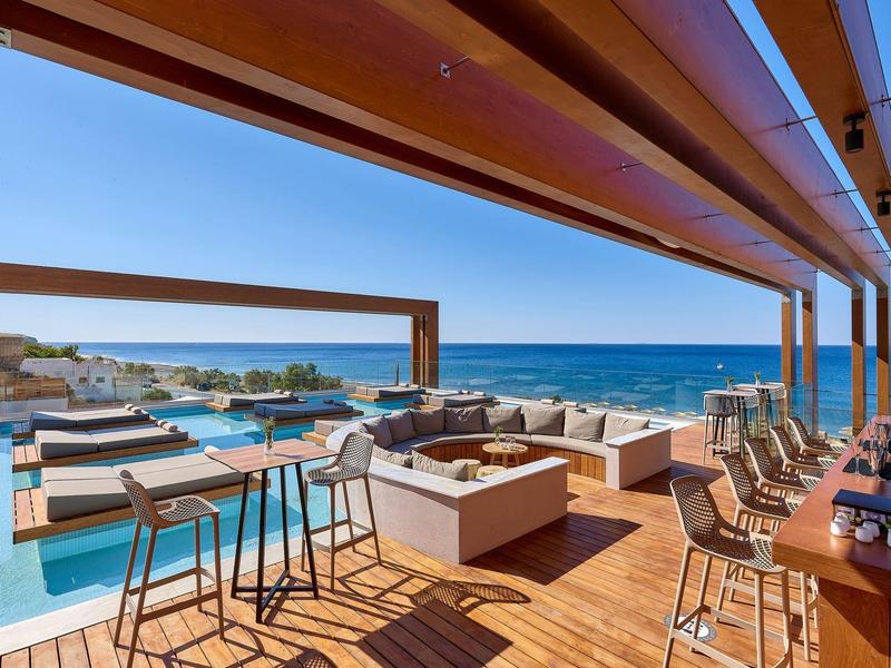 Střešní lounge s barem, posezením a bazény s výhledem na oceán pod jasnou modrou oblohou.