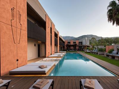 Area piscina moderna con terrazza solarium, lettini, palme, edificio dell'hotel e montagne