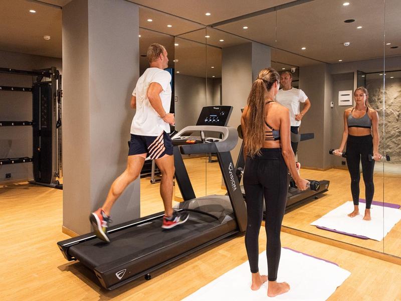 Deux personnes dans une salle de sport : un homme sur le tapis roulant et une femme devant un miroir.