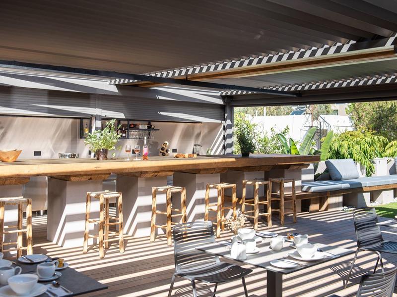 Espace de restauration moderne ouvert avec bar en bois, tabourets et sièges extérieurs avec canapés ensoleillés.