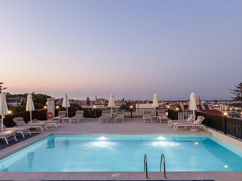 Verlicht hotelzwembad met ligstoelen en zitplaatsen op dakterras bij zonsondergang.