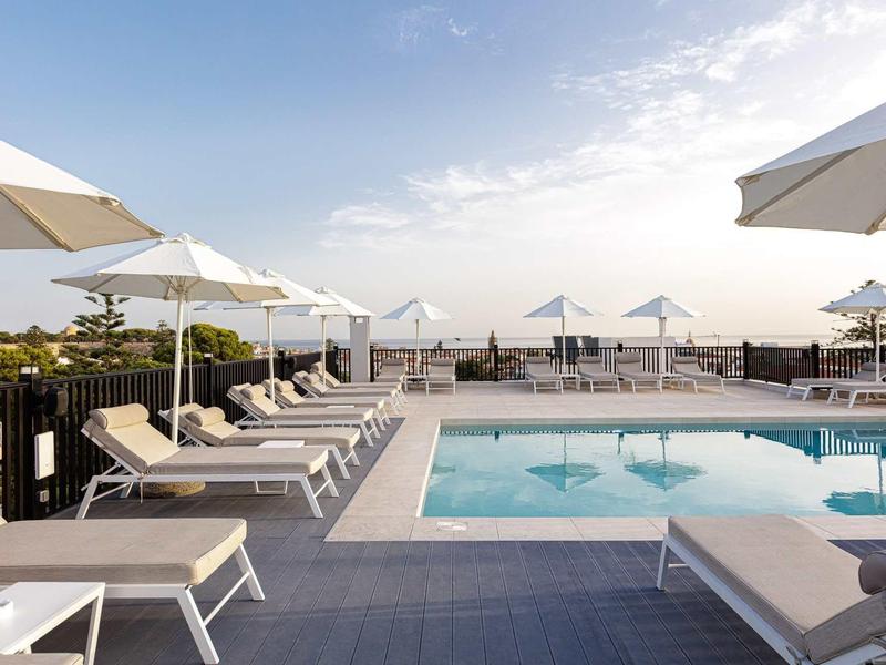 Moderne buitenzwembad met ligstoelen en parasols op een hotelterras met uitzicht op zee.