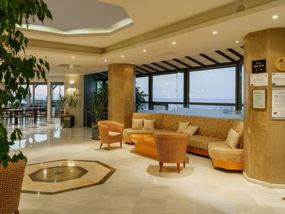 Hotel lobby with beige sofa, chairs, large windows, and polished floor with plant.
