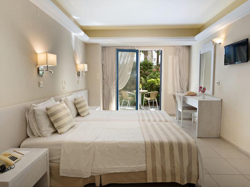 Bright hotel room with double bed, striped linens, desk, and balcony with garden view.