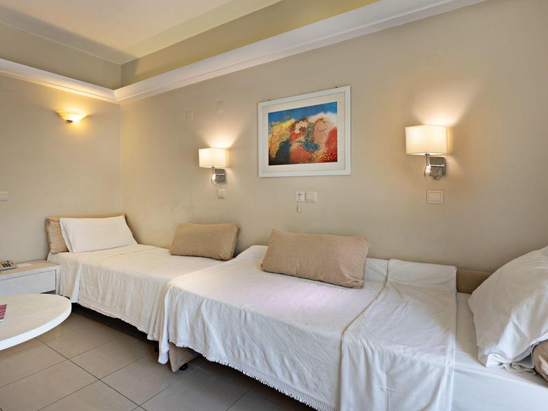 Simple hotel room with two single beds, beige walls, soft lighting, and a framed painting.