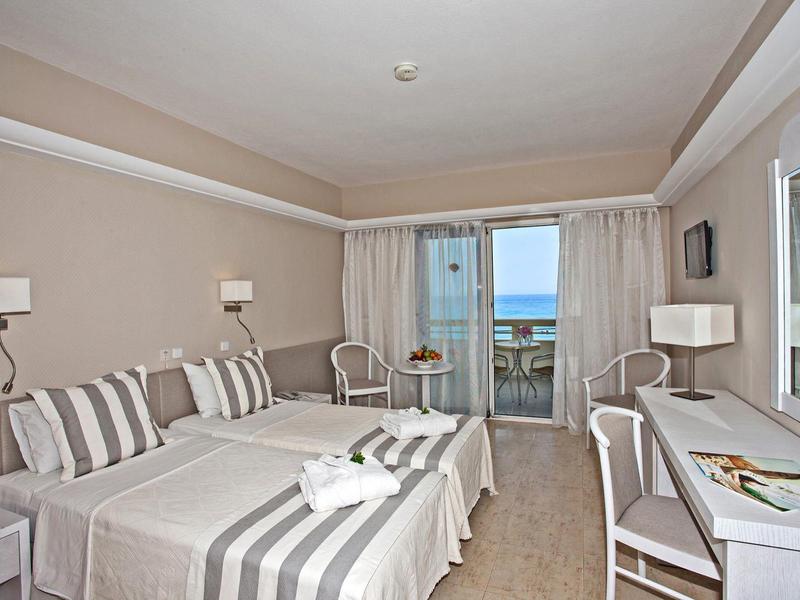 Bright hotel room with twin beds, striped bedding, a desk, and a balcony with sea view.