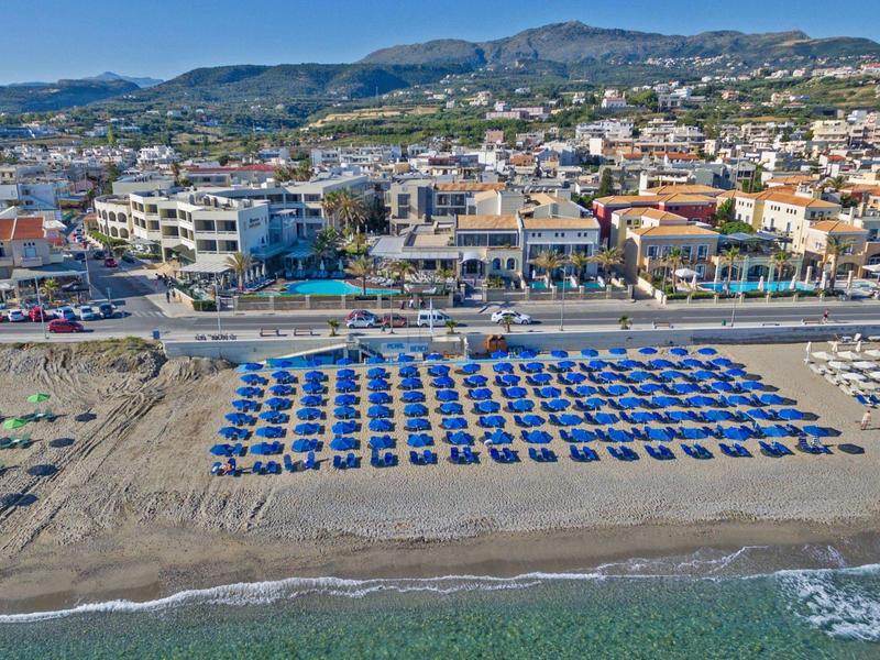 Beach with rows of blue umbrellas and sunbeds near a coastal town under clear sky.