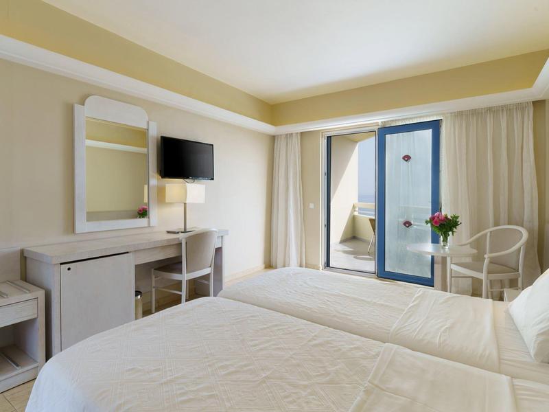 Hotel room with two beds, desk, TV, and a balcony with chairs and flowers.