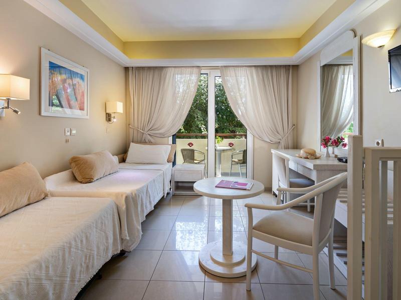 Bright hotel room with two beds, a table, chairs, TV, and a balcony with garden view.